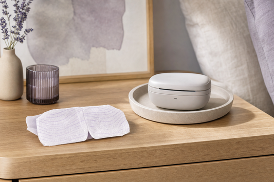 Folded unbranded disposable eye mask beside a closed rechargeable eye-care device on a wooden nightstand in soft daylight.